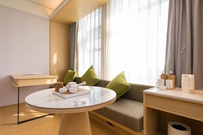 Et moderne hotelværelse med et rundt marmorbord, en sofa med grønne puder, et træsidebord med toiletartikler, og et vindue med lette gardiner.