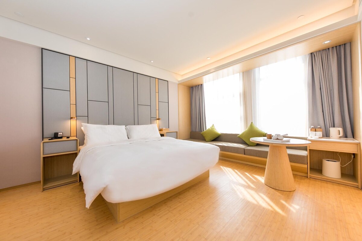 A modern hotel room with a large bed, a seating area with a table, and a window with curtains.