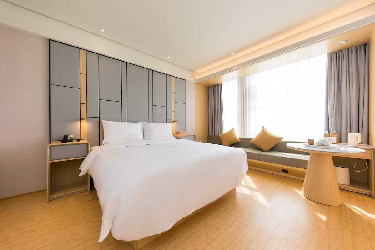 A modern hotel room with a large bed, bedside tables, a round breakfast table, and a seating area with a sofa and cushions.