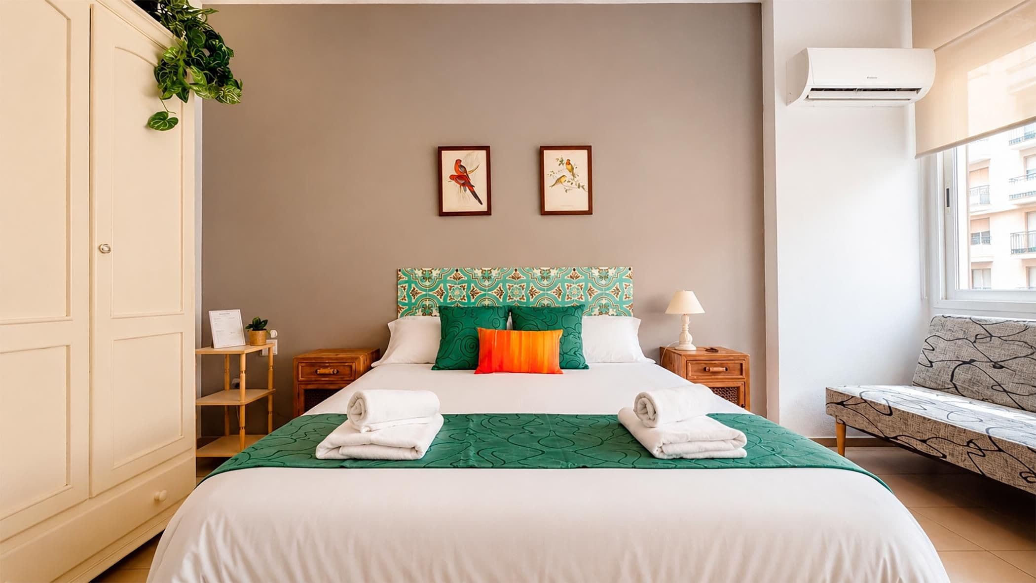 Ein Hotelzimmer mit einem Bett, zwei Nachttischen, einer Sitzecke und einem Fenster. Eine Klimaanlage ist ebenfalls vorhanden.