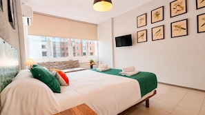A hotel room with a bed, a TV, a window with a view of buildings, and framed pictures on the wall.