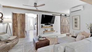 Amplia sala de estar con un sofá grande, una mesa de centro y un televisor montado en la pared. Se ve una puerta de madera que da a otra habitación y un ventilador de techo.