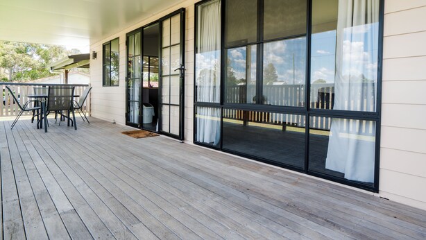 Un espacio al aire libre techado con piso de madera, una mesa con sillas y grandes puertas corredizas de vidrio que dan a otra terraza.