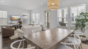 Un salon moderne avec un coin repas, des meubles blancs et une grande fenêtre équipée de stores.
