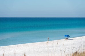 Ein Strand mit blauem Meer, sandigem Ufer und einer Person unter einem blauen Sonnenschirm.