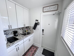 Cocina compacta con armarios blancos, encimera negra, cafetera y una puerta con picaporte.