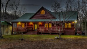 Une cabane en rondins avec un toit végétalisé, de nombreuses fenêtres et une véranda illuminée.
