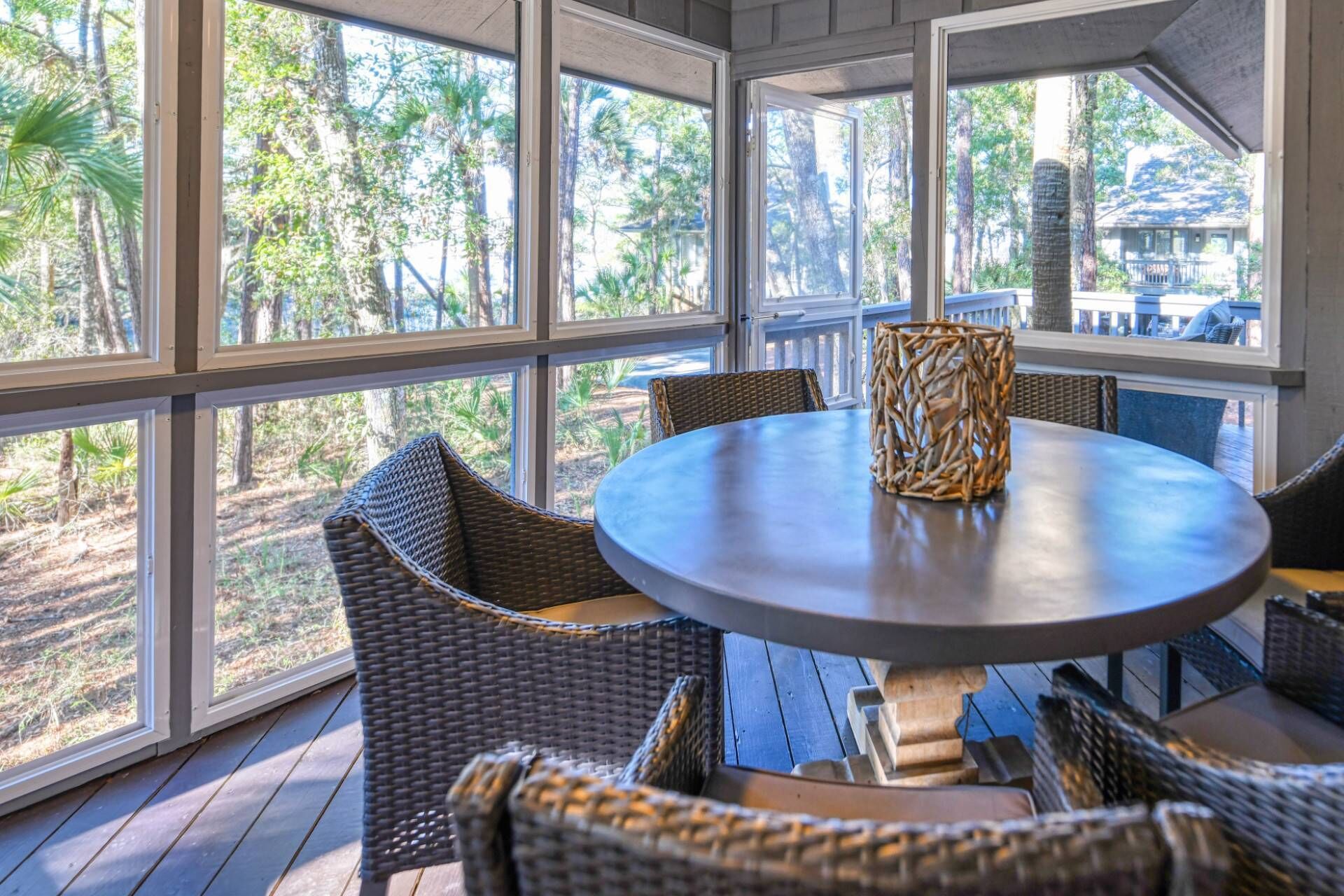 Una mesa redonda con sillas de mimbre y un centro de mesa tejido, ubicada en una habitación con grandes ventanales con vistas a un área boscosa.
