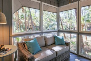 A cozy room with a wicker sofa, a small round table, and large windows offering a view of a forested area.