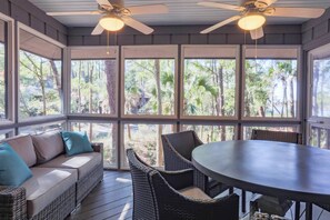 A covered outdoor seating area with a sofa, chairs, and a table, featuring large windows and ceiling fans.