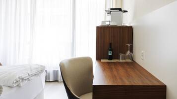 A hotel room with a bed, a wooden desk, a chair, a bottle, and a coffee maker.