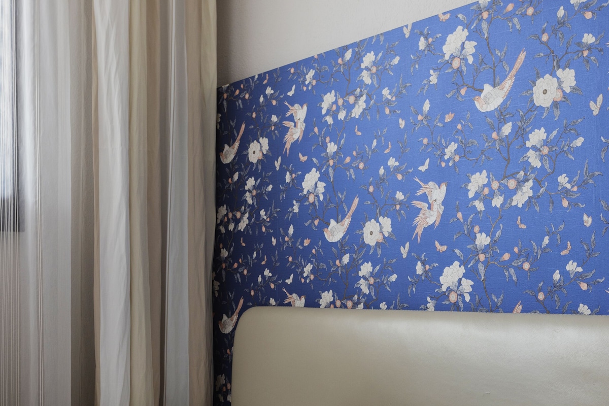 A bed with white bedding, a blue floral headboard, and sheer curtains.