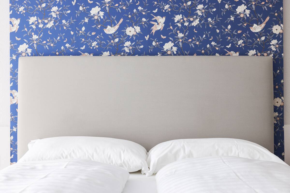 A bed with white bedding and a plain headboard against a blue floral wallpaper.