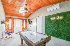 Um quarto com mesa de pebolim, ventilador de teto, aparelho de ar condicionado, televisão e um letreiro com a frase 'Hello Chico' em uma parede verde.