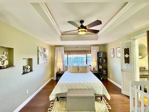 Um quarto com cama, ventilador de teto, janela com cortinas, mesa de cabeceira e criado-mudo.