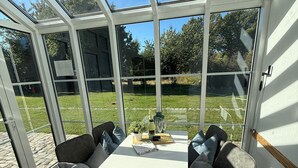 A conservatory with a glass roof, a white table, grey sofas, and a view of a grassy area.