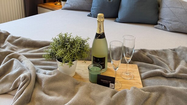 A neatly made bed with a champagne bottle, glasses, and a card on a wooden tray, a potted plant, and a painting on the wall.