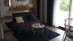 Une pièce confortable avec un lit, une table basse ronde, une chaise, une étagère ornée d’objets décoratifs et une fenêtre avec des rideaux.