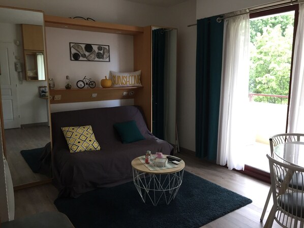 A cozy room with a bed, a round coffee table, a chair, a shelf with decorative items, and a window with curtains. - Quiet apartment in the heart of La Rochelle, 5 minutes from the beach (La Rochelle)