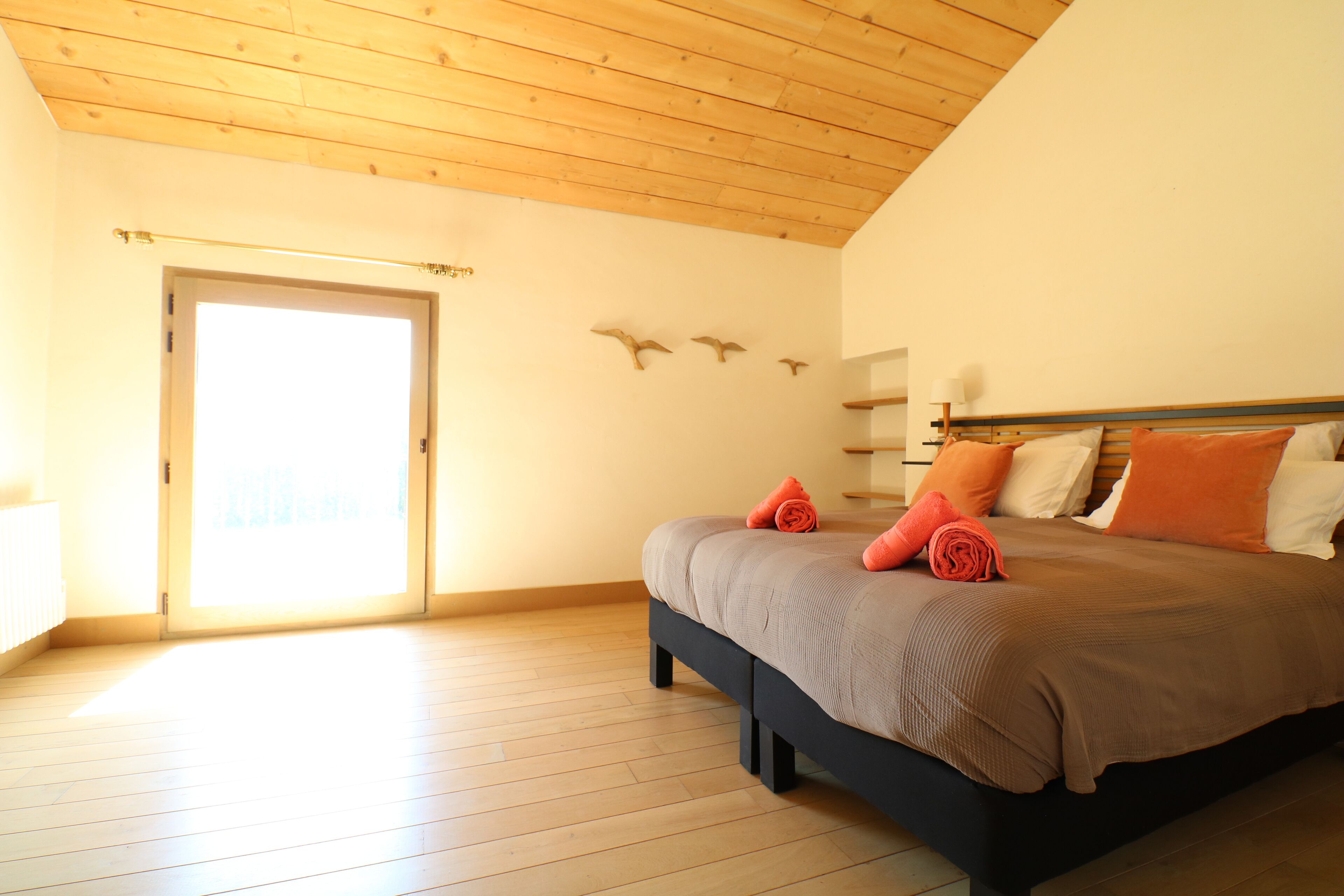 Un dormitorio con suelo de madera, una cama con colcha marrón y una puerta que da a una habitación luminosa.