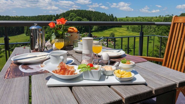Een ontbijttafel op een balkon met een mooi uitzicht, inclusief een metalen bak, een vaas met bloemen en diverse ontbijtartikelen.