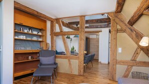 Een knus vertrek met zichtbare houten balken, een grijze fauteuil, een klein rond tafeltje, en een keukenhoek met planken en een hangende plant.