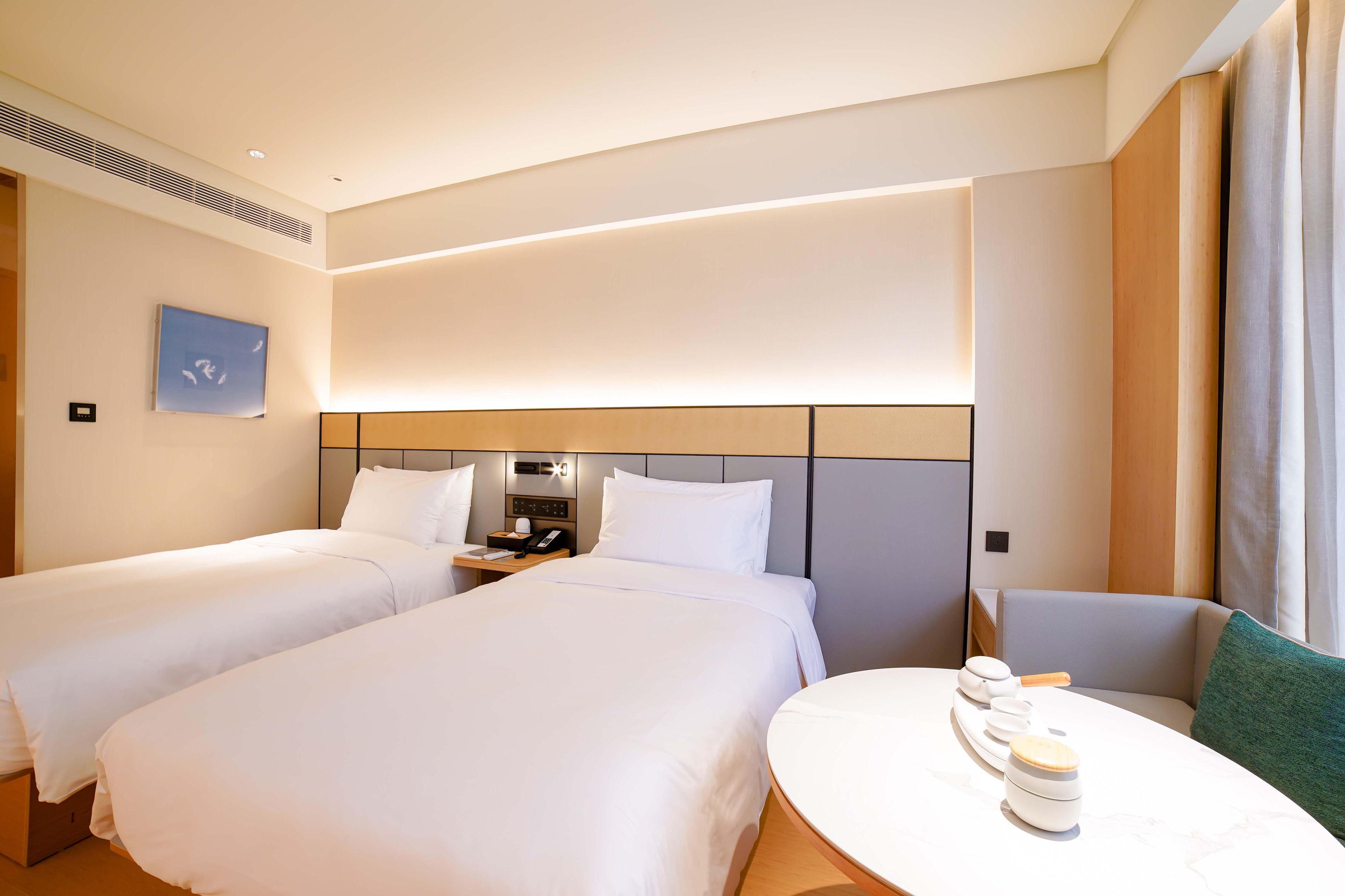A hotel room with two beds, a bedside table with a phone and a lamp, a small round table with a kettle, and a painting on the wall.