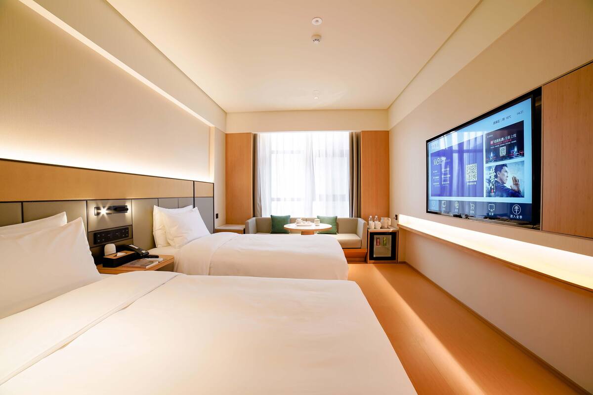 A hotel room with two beds, a TV mounted on the wall, a small table, and a window with curtains.