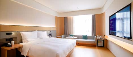Quarto de hotel moderno com uma cama grande, uma mesa de cabeceira com telefone, uma pequena área de estar com mesa e uma televisão fixada na parede.