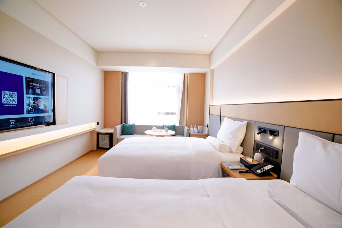 A hotel room with two beds, a TV on the wall, a small table with a lamp, and a window with curtains.