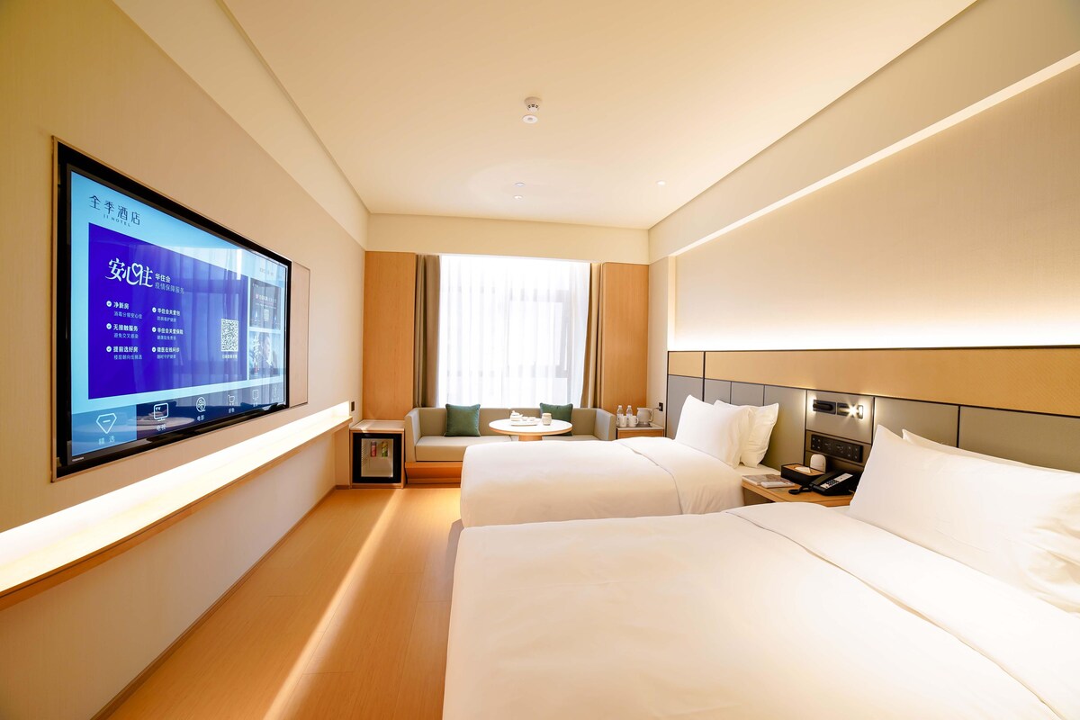 A hotel room with two beds, a TV mounted on the wall, a small table, and a sofa.