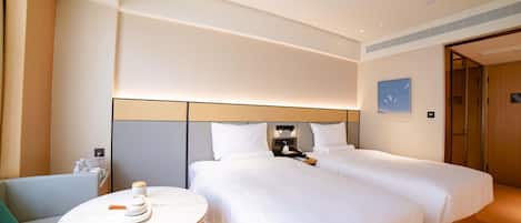 A hotel room with two beds, a small round table, a chair, a bedside table with a lamp, and a wall-mounted control panel.