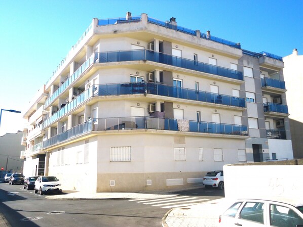 Un immeuble résidentiel à plusieurs étages, doté de balcons, donnant sur la rue et avec des voitures garées devant.