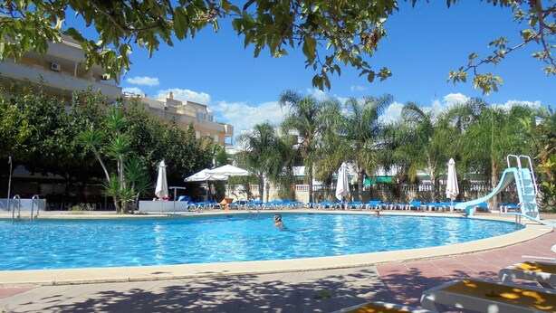 Una piscina con sillas de descanso y sombrillas, rodeada de palmeras y un edificio al fondo.