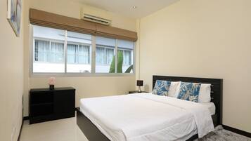 A bedroom with a bed, a nightstand, a lamp, a window with blinds, and an air conditioning unit.