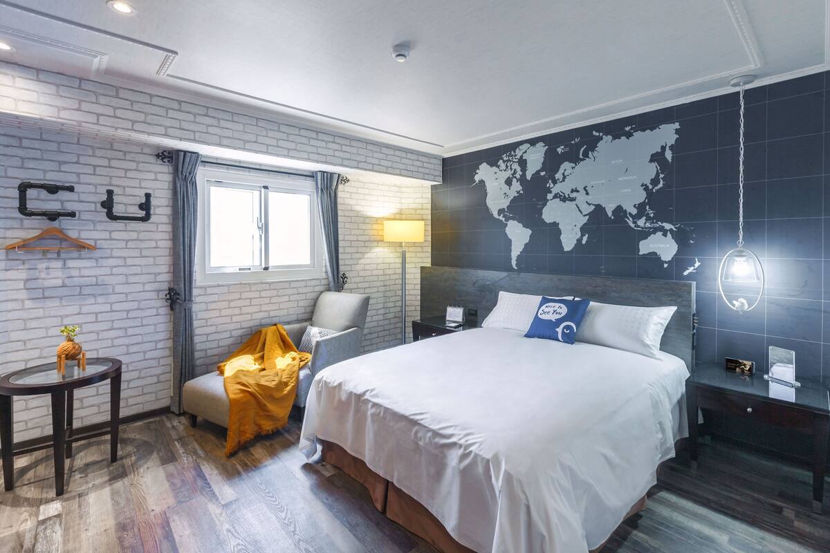 A hotel room with a large bed, a world map on the wall, a small table with a lamp, and a chair with a yellow blanket.