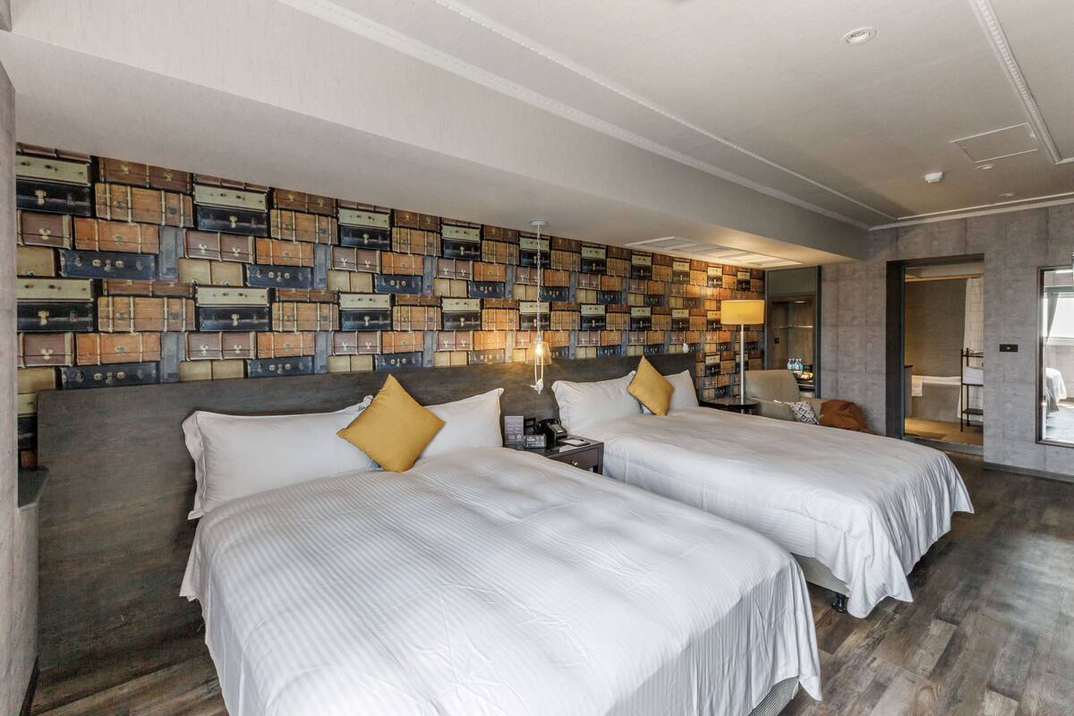 A hotel room with two beds, a wall of stacked suitcases, a wooden floor, and a modern bathroom visible through an open door.