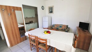 Un coin repas compact avec une table dressée pour deux personnes, une petite kitchenette à l’arrière-plan, un téléviseur posé sur un meuble en bois et une horloge au mur.