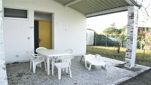 Une terrasse couverte avec une table blanche et quatre chaises, une porte jaune et un pilier en briques.