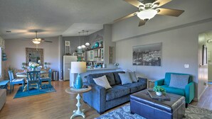 Een ruime woonkamer met een grijze bank, een blauwgroene fauteuil en een salontafel. Er is een eethoek met een tafel en stoelen, en een keuken met kasten en een koelkast.