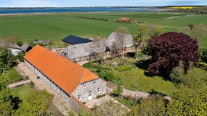Ein Steinhaus mit rotem Dach, umgeben von grünen Feldern und einem großen Gewässer im Hintergrund.