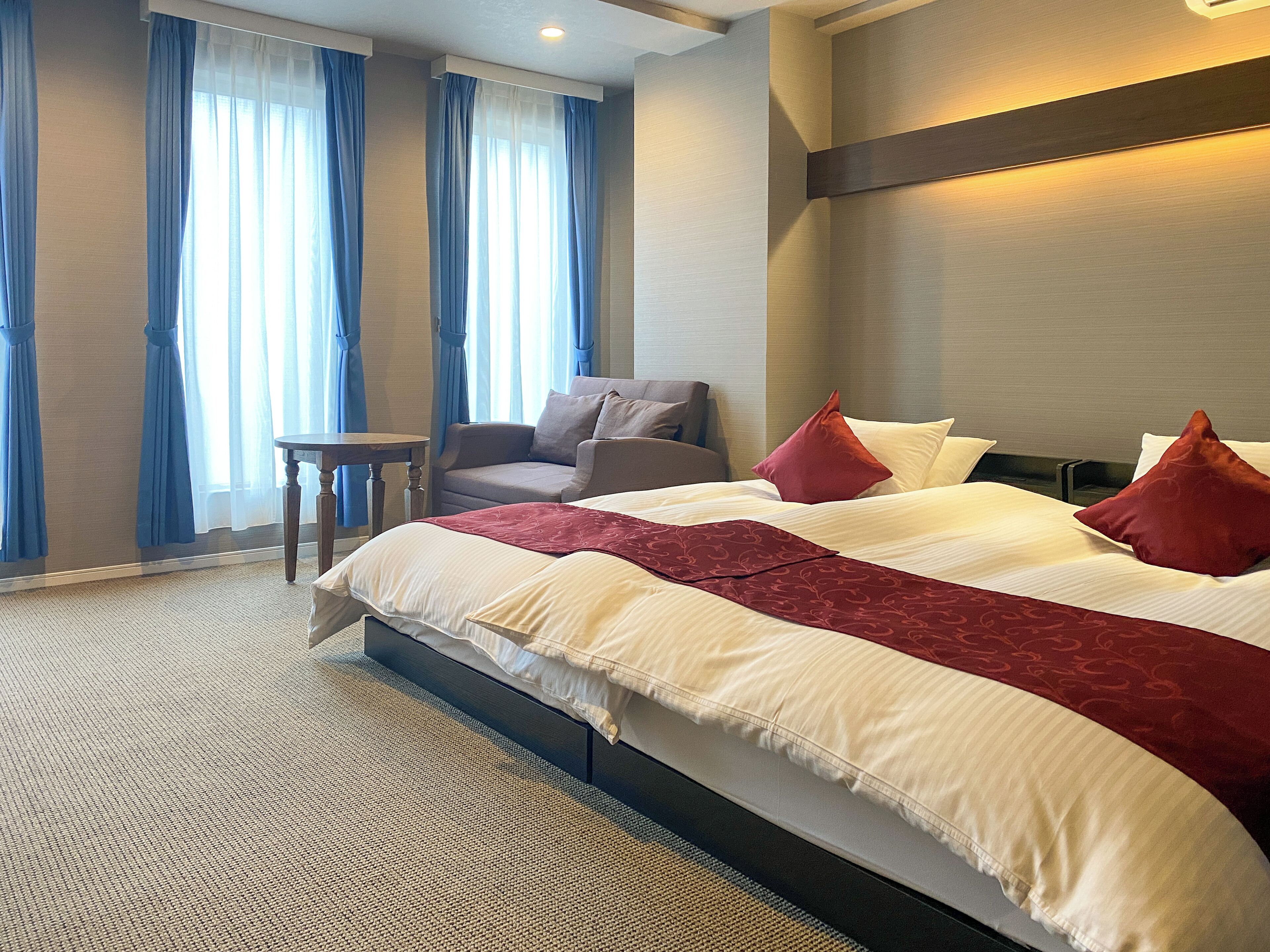 A hotel room with a large bed, a chair, a small table, and a view of the outdoors through the window.