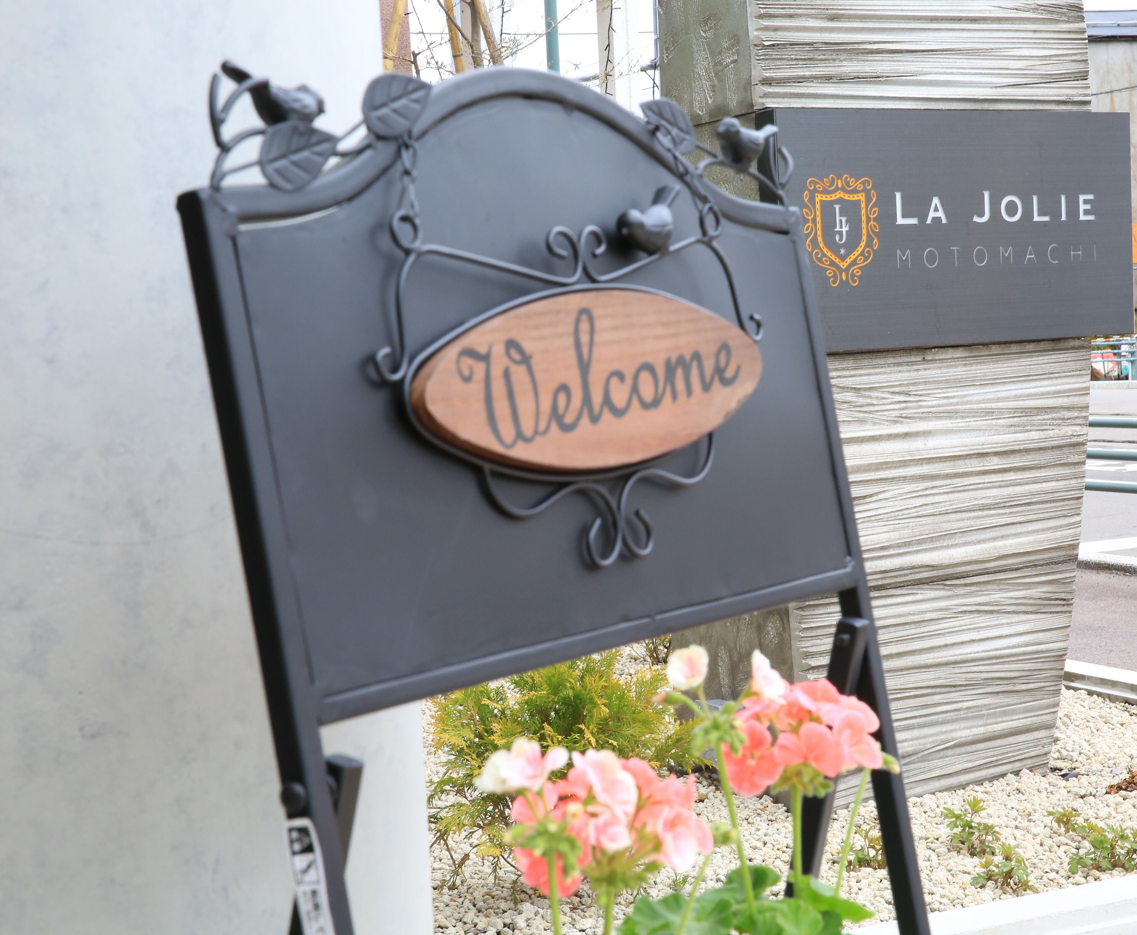 A welcome sign with a wooden plaque and metal frame, placed in front of a building with a sign reading "LA JOLIE MOTOMACHI".