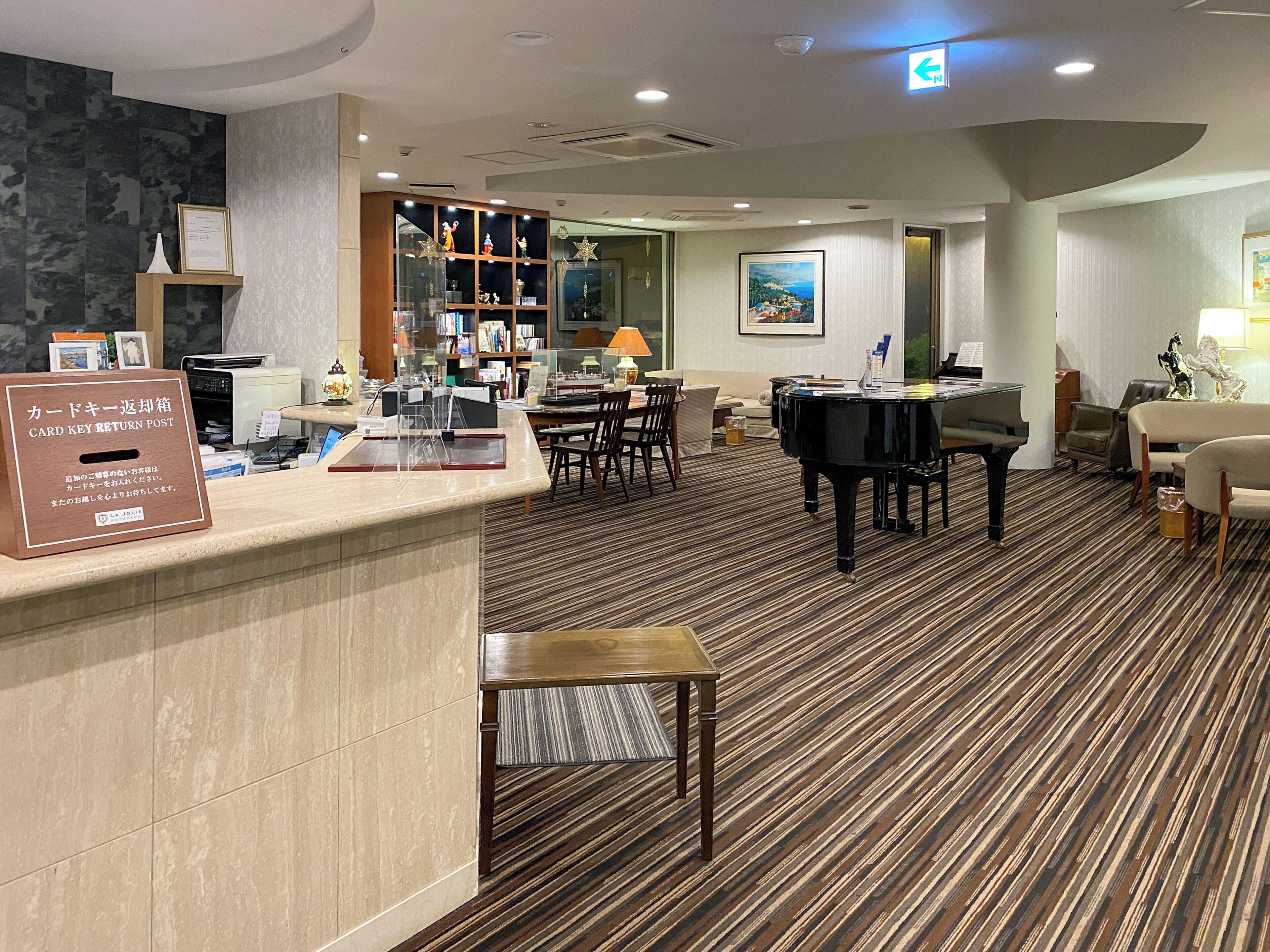Un hall d’hôtel comprenant un comptoir d’accueil, un piano, un espace salon et une bibliothèque contenant divers objets.