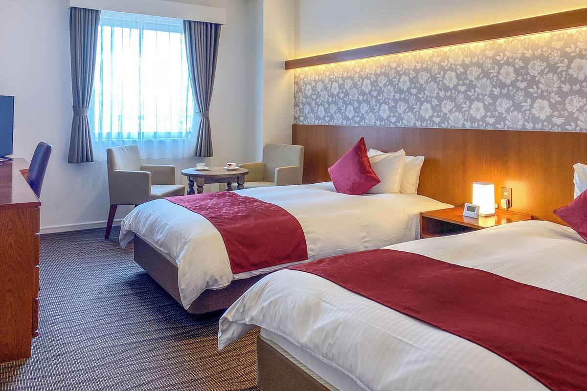 Een hotelkamer met twee bedden, een bureau, een stoel, een bijzettafeltje en een raam met gordijnen.