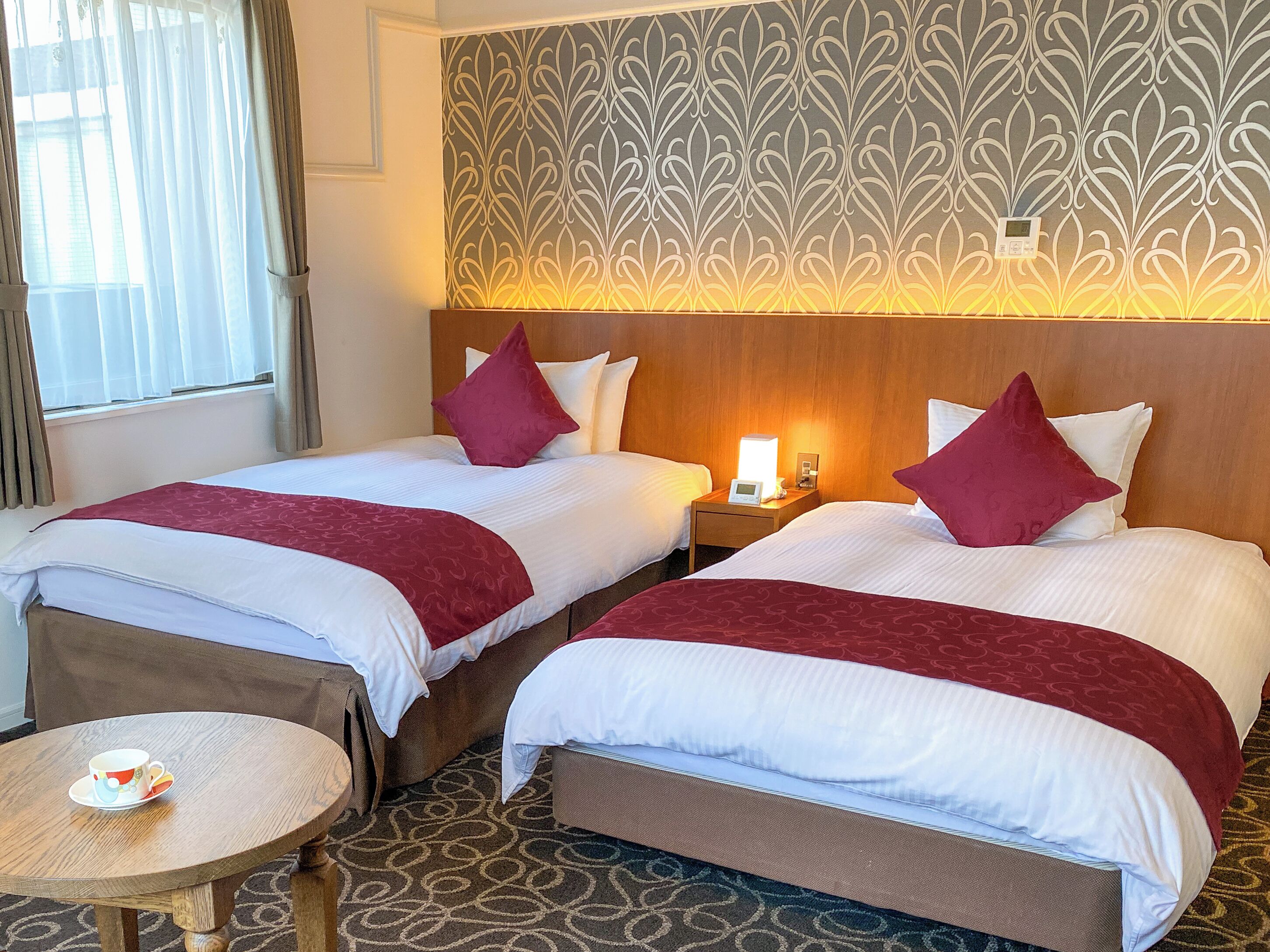A hotel room with two beds, a bedside table, a lamp, and a small round table with a cup.