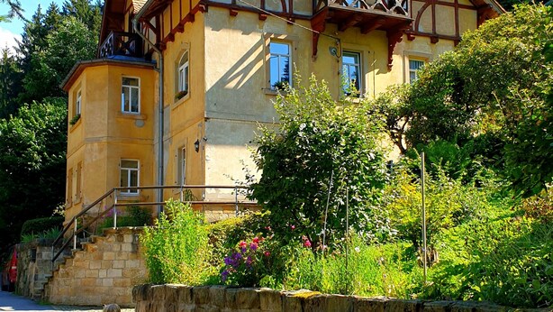 A traditional half-timbered house with a balcony, surrounded by lush greenery and a stone wall. - Apartment Ostrau | Villa Falkenstein (Bad Schandau)