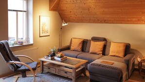 A cozy living room with a wooden ceiling, a sofa with cushions, a coffee table with books and a vase, and a chair. - Apartment Ostrau | Villa Falkenstein (Bad Schandau)