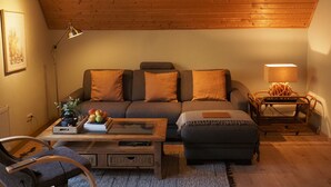 A cozy living room with a wooden ceiling, a sofa with matching cushions, a coffee table with books and fruit, and a lamp on a side table. - Apartment Ostrau | Villa Falkenstein (Bad Schandau)