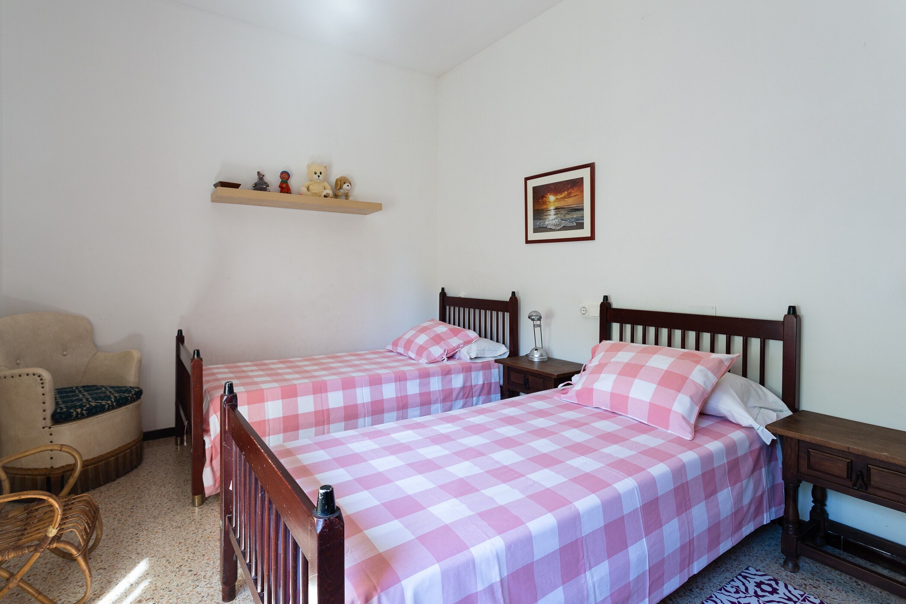 Un dormitorio con dos camas, una silla, una mesita de noche, una estantería con adornos y un cuadro en la pared.